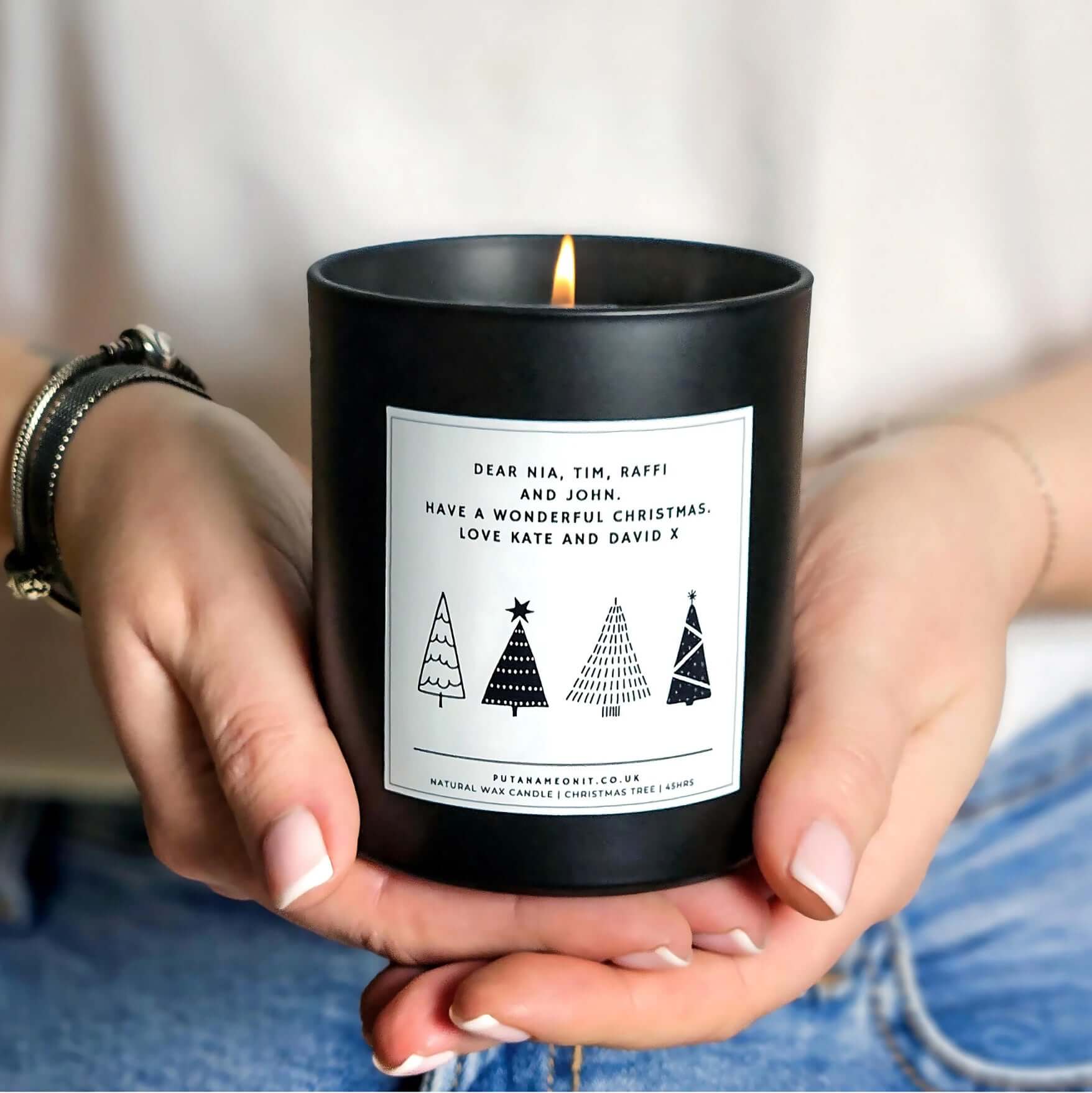 Hands holding a personalised Christmas candle in a matte black jar, featuring a monochrome minimalist Christmas tree design on a white label with customisable names and message