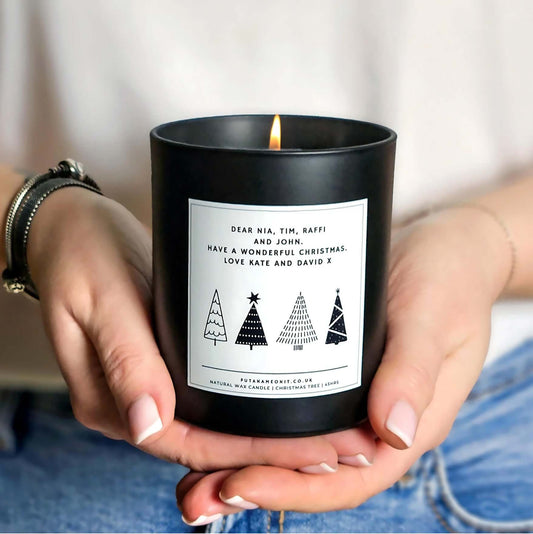 Hands holding a personalised Christmas candle in a matte black jar, featuring a monochrome minimalist Christmas tree design on a white label with customisable names and message