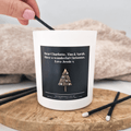 Hand lighting a personalised Christmas candle with a sleek black label featuring a gold minimalist Christmas tree design and a custom festive message. The candle is presented in a white glass jar on a wooden surface with black candle matches.