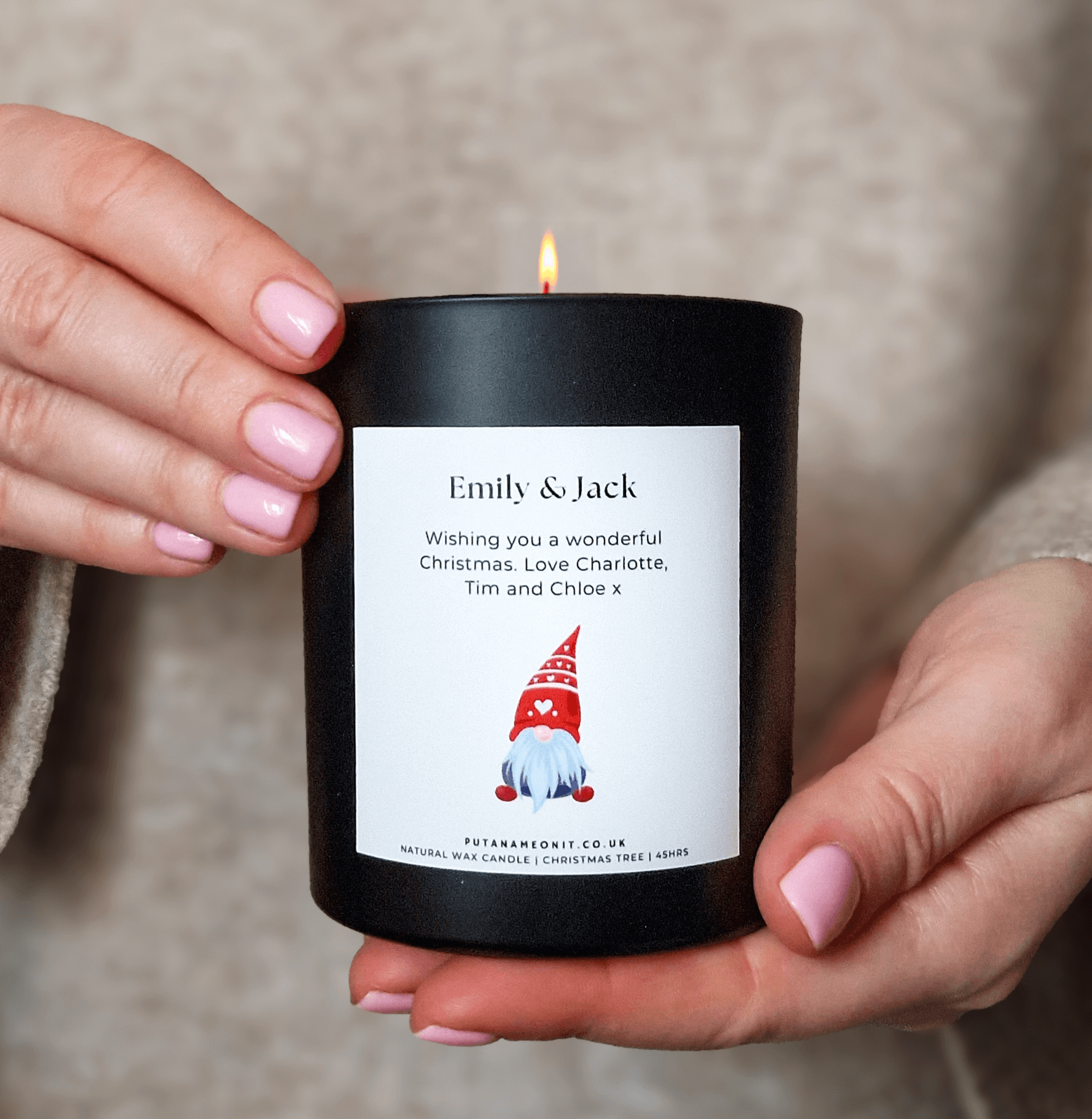 Person holding a personalised Christmas candle in a black jar with a white label featuring a red festive gnome design, custom names, and a holiday message. The candle emits a warm glow, perfect for gifting or festive décor